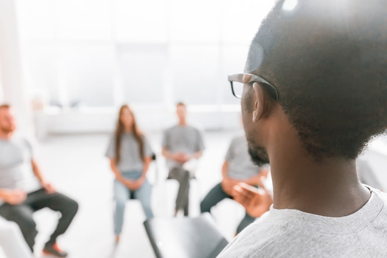 Blurred people sat on chairs with on man standing, not blurred at the front